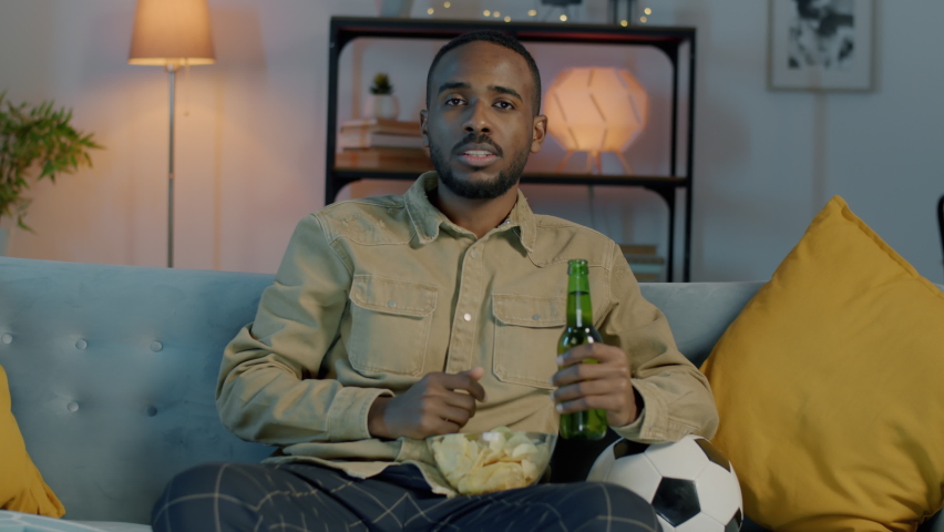 Happy African American man watching football game on TV eating crisps and cheering at home at night. Entertainment and modern lifestyle concept. Royalty-Free Stock Footage #1091891817