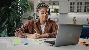 Online education. Young cute black lady student wearing headset with mic video chatting with tutor via laptop, writing notes in copybook, zoom in portrait, slow motion