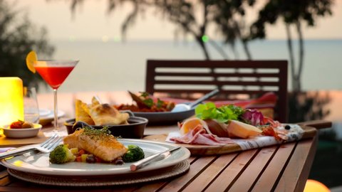 Romantic dinner served on wooden table at sunset with sea, horizon, sky background. Fine dining restaurant at luxury hotel or resort. Grilled salmon with broccoli on ceramic plate, cocktail, cold cut.
