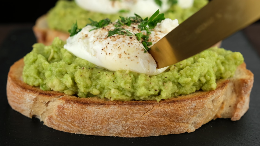 Cutting poached egg with runny egg yolk over bread toast with mashed avocado spread. Avocado toast with poached egg close up. Healthy breakfast or lunch food. Royalty-Free Stock Footage #1091901373