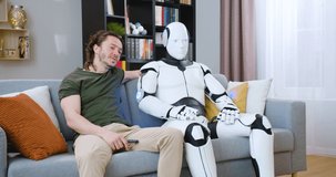 Smiling young guy in casual wear sitting on cozy sofa and talking with android friend while watching TV together at home. Concept of artificial intelligence and leisure time spending.