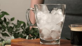 4k video, pouring brewing coffee into glass full of ice is served with cookies on wooden table. Making cold coffee drink in slow motion. 