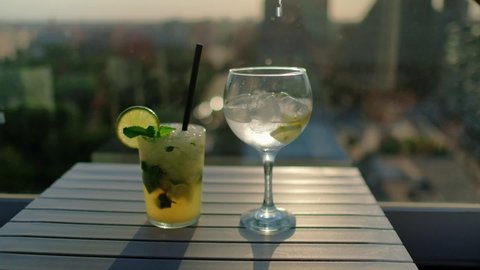 Tropical cocktail decorated with fresh lime and mint leaves. Mojito and gin tonic drinks. Beautiful sunset on the background. Vacation summer vibe. Full HD slow motion video. Sunset on the background.