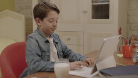 Little boy is drinking milk while doing his lessons on the tablet computer. He takes computer lessons, plays games, and watches videos while drinking his milk at his desk in his room. Milk is health.