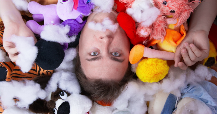 Plump man lies on heap of different plush toys. Scared man spits stuffing from soft animals out of mouth. Mess at home created by dogs close upper view