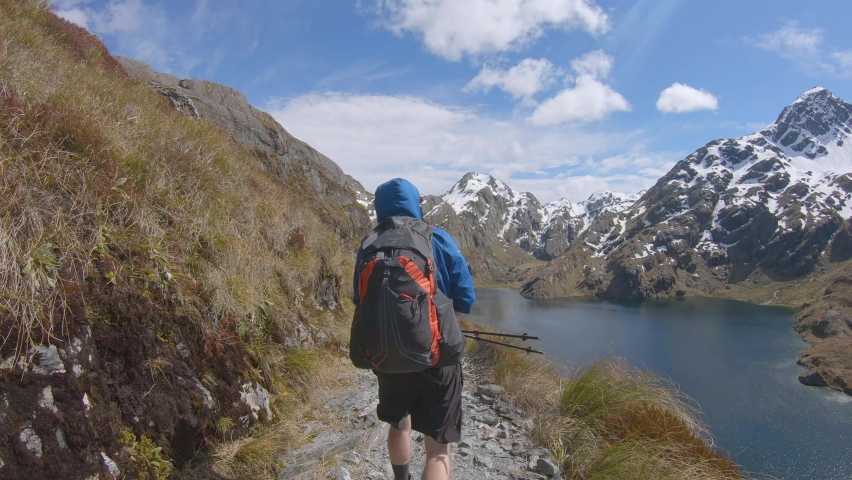 Follow, hiker walks above alpine lake, snow capped mountain landscape, Routeburn Track New Zealand