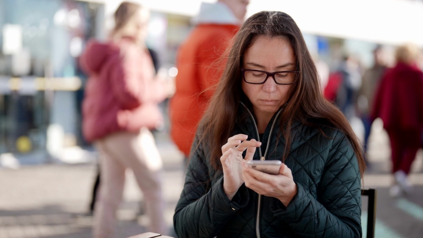 A close-up portrait of an adult woman with glasses sitting at a table in an outdoor cafe, writing a message on a smartphone. A business woman using a mobile phone outdoors in an urban background.