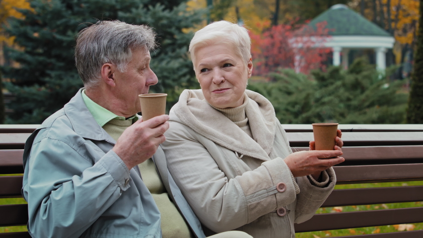 Married couple elderly caucasian family two 60s people happy grandparents pensioners sit in autumn park drinking hot coffee tea outdoors resting together relaxing romantic relations enjoying leisure Royalty-Free Stock Footage #1091938283