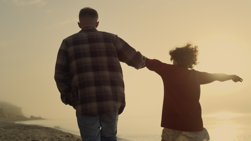 Back view affectionate couple holding hands on beach. Cheerful woman and man walking along sea shore at sunset. Playful girl and guy enjoying romantic time. Young hipsters having fun. Love concept  Royalty-Free Stock Footage #1091939225