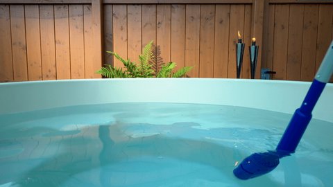 A battery operated pool vacuum is used to clean a hot tub. Set against a backdrop of a beautiful wooden fence, a healthy green fern illuminated by a streak of sunlight and two chrome garden torches.