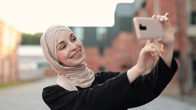 Portrait of smiling happy beautiful muslim woman relaxing using digital smartphone. Young muslim girl looking at screen speaking during online video call on social media on city.