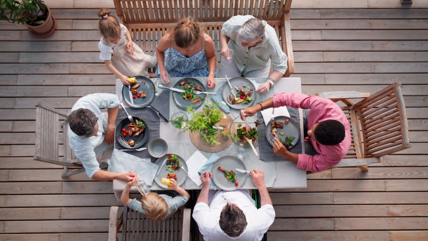 Top view of 3 generations family eating at barbecue party dinner on patio, people sitting at table on patio with grill.