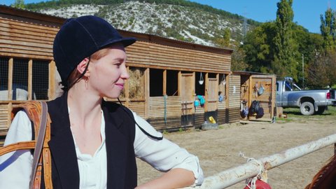 Happy female equestrienne with harness on shoulder looks at horse standing nearby. Rider caucasian girl is near stabling on paddock fenced area. Jockette smiling, resting next to palfrey outdoor