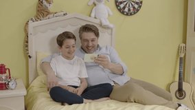 Image of cheerful father using smartphone and lying in bed with little son.Smiling father and cute little boy lying in bed and playing video game on mobile phones while having fun together at home.
