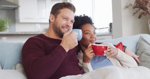 Video of happy diverse couple embracing and drinking coffee on sofa. Love, relationship and spending quality time together at home.