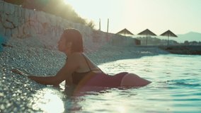 Fashion video of a girl lying on the beach and the waves crashing against her. Beautiful woman in a bathing suit relaxing by the sea at sunset.