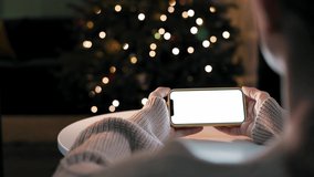 Girl holds smartphone in hands with white blank screen at home against the background of festive New Year's lights on the Christmas tree. Watching movie, news, social media, video call online