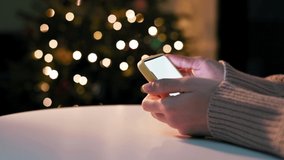 Girl holds smartphone in hands with white blank screen at home against the background of festive New Year's lights on the Christmas tree. Watching movie, news, social media, video call online