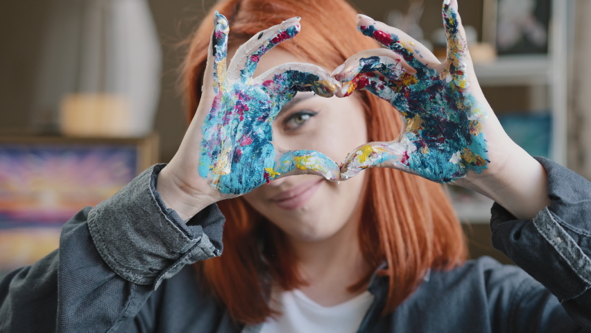 Portrait red-haired girl talented artist female painter stands posing in workshop shows gesture of kindness inspiration love for art sign heart shape with dirty hands in oil acrylic paints smiling Royalty-Free Stock Footage #1091980293