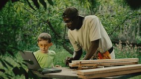 African American carpenter and schooler watch video guide of making table from wooden planks via laptop. Specialist and boy enjoy woodwork in garden