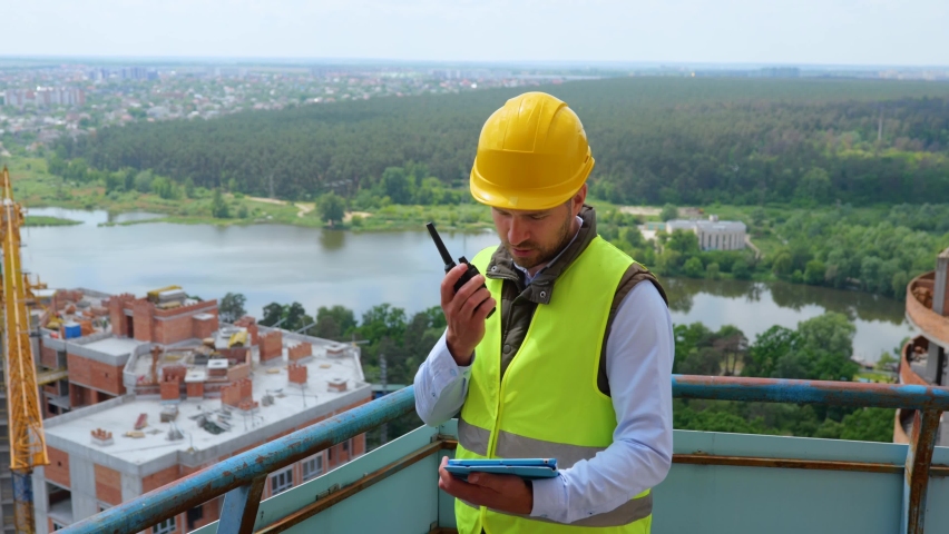 Portrait of Caucasian handsome man builder manager in uniform and helmet looking at digital tablet and talking in walkie-talkie while standing on construction site. Work, business