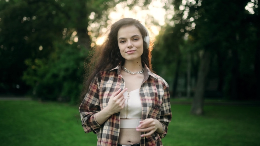 Cinematic shot of young happy woman is having fun to listening to the music with earphones and dancing in green city park at sunset
