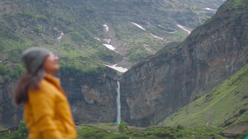 Rack focus shot of Sissu waterfall with a female Indian tourist standing in front of it at Lahaul and Spiti district, Himachal pradesh, India. Focus changes from waterfall to female traveler.  | Shutterstock HD Video #1091985783