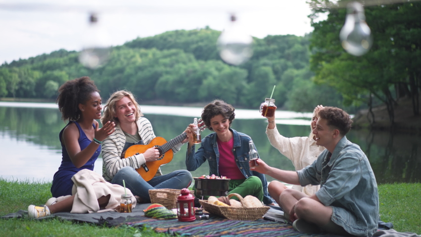 Group of friends having fun on picnic near a lake, sitting on blanket eating and playing guitar.