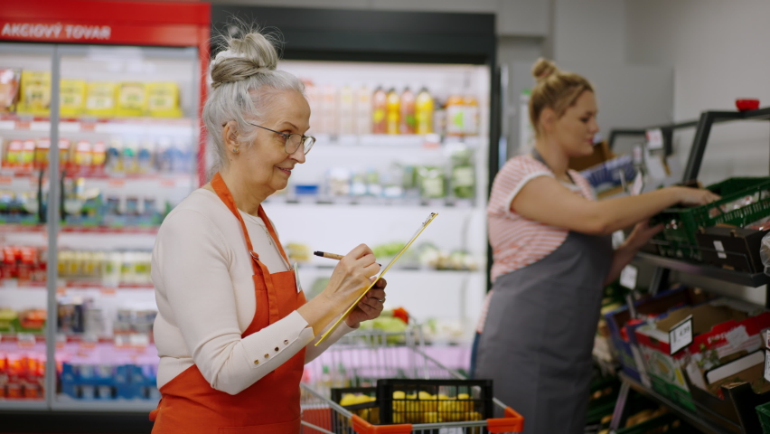 Confident senior shop assistant in supermarket in vegetable shell, in bakcground is her colleague filling stock.