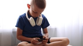 Child's dependence on a mobile device theme. Child with headphones on neck is sitting on table and uncontrollably playing online games on smartphone. virtual life. children's addiction to online games