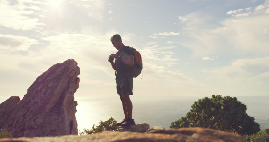 Fit active man hiking, standing on a mountain peak with binoculars and a backpack during a hike and enjoying the landscape view in front of him. Male hiker standing on a rock looking around at nature
