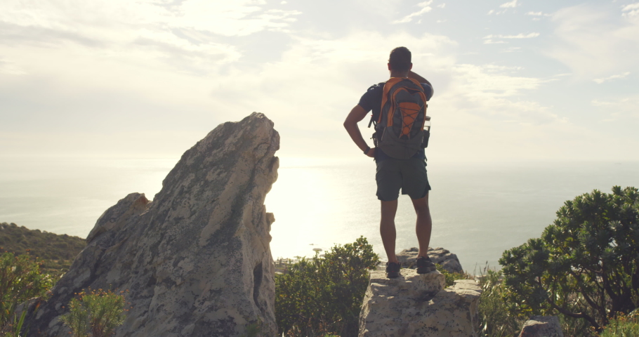 Rear view of fit active man standing with a backpack during a hike and enjoying the coastal landscape in front of him with seascape and coast. Male hiker standing on a rock looking at a nature view