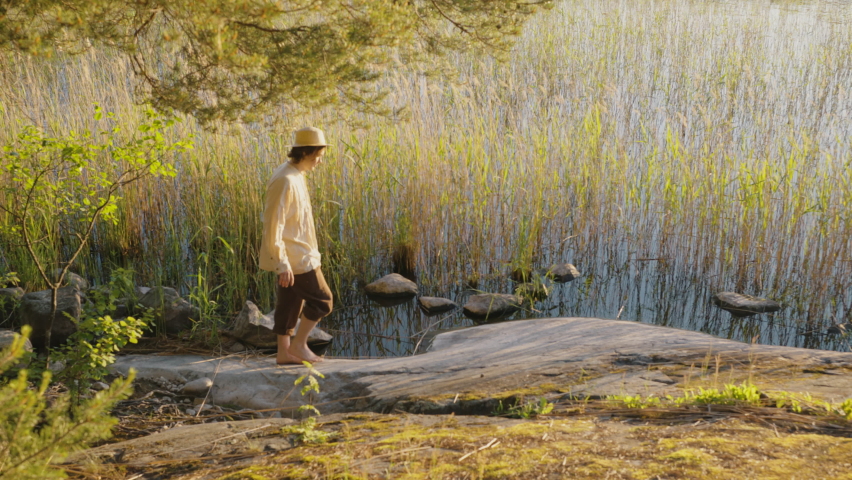 Young man walks barefoot near lake water with bulrush and rocks on sunny day. Man wearing white hat and shirt enjoys vacation in nature