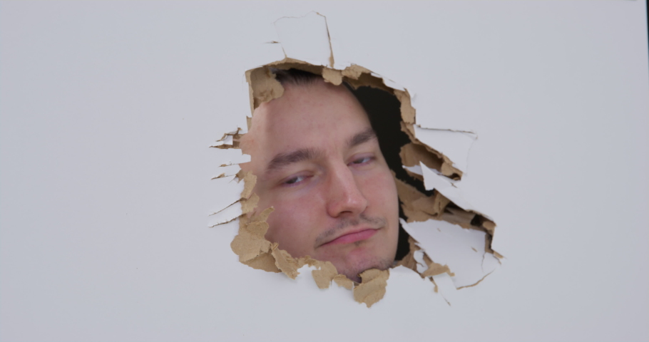 Young smiling man looks out of a punched hole in a plasterboard door, and then disappears, front view. Person is fooling around during the renovation of apartment