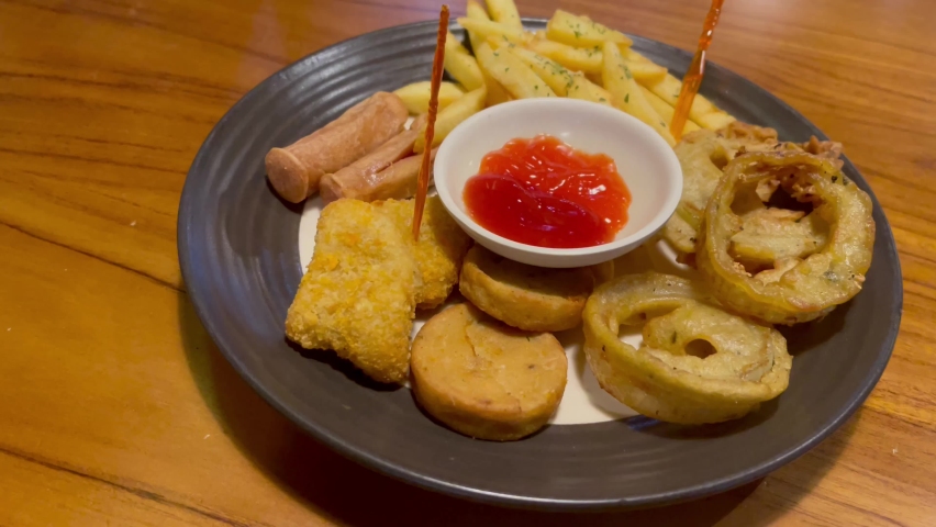 A mix platter menu at a cafe or restaurant consisting of onion rings, potato sticks, nuggets, chicken or beef sausage with tomato sauce and small plastic forks on a white plate on a wooden table.