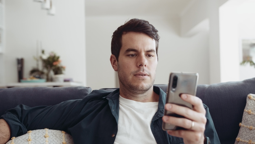 Caucasian man sitting on couch at home and is doing business on his mobile phone. He becomes frustrated, sighs and puts his mobile phone down. 