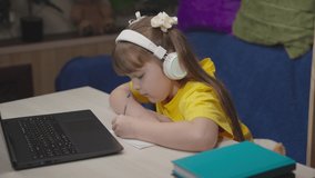 school girl headphones does homework laptop while sitting table room. children classes online home. student concept. girl daughter writes with pen notebook. remote school home learning child. school