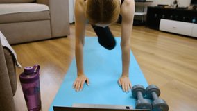 A young girl is engaged in fitness at home. The girl found an exercise on a laptop and is doing it. The girl is engaged on a sports mat. Slow motion.