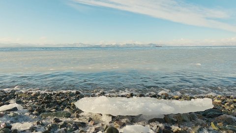 Seascape in winter. Bay with surf and ships on the horizon. Small ice floes float in the sea. The beach is pebbly and covered with snow. Kamchatka. Russia. Pacific Ocean.