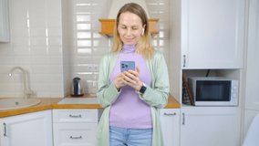 Cheerful Ukrainain woman typing message in messenger app on mobile phone.Happy white blonde in 40s communicating online with modern smartphone and listening to music in wireless headphones in 4k video