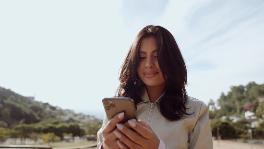 Beautiful young Latin woman using smartphone standing on city street. Portrait of Latina smiling female using mobile phone. Cinematic 4K Royalty-Free Stock Footage #1092019523