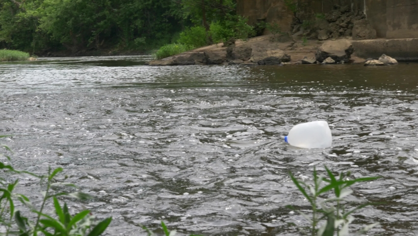milk jug floats down river slowly behind foliage towards old concrete bridge on a cloudy day slow motion Royalty-Free Stock Footage #1092020843