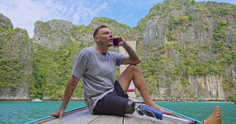 Young caucasian male traveler with artificial bionic leg prosthesis sitting on the front of long tail boat uses his phone to chat with people online and share the breathtaking scenery in background.