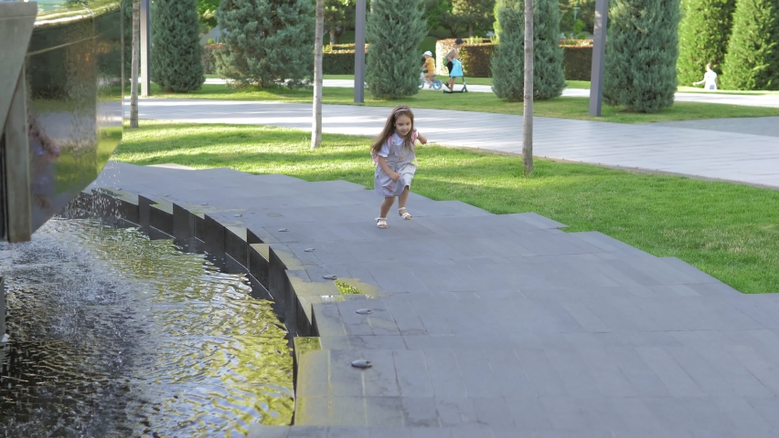 A little girl in a dress runs along the fountain to her parent. Happy childhood, lifestyle. A Caucasian child runs along a path on a bright sunny day in a public park. Children
