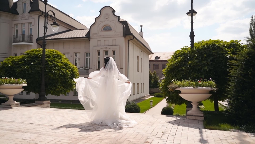 Pretty bride in long airy veil, elegant white dress spins with fabric fluttering in air on wedding day in luxury palace garden. Playful attractive woman posing on camera swirling in fabulous gown. Royalty-Free Stock Footage #1092049387