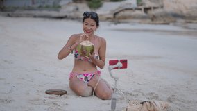 Asian Thai girl recording live stream sitting on a beach and drinking coconut water at sunset
