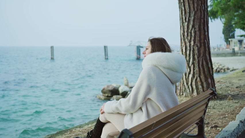 Young lady in white furry coat sits on bench looking at boats sailing on lake Garda. Big waves on water surface caused by wind on autumn day