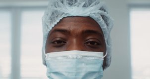 Close-up of the face of an African-American doctor wearing a medical mask and a disposable cap on his head. Smiling eyes of a doctor. 4k video, red komodo