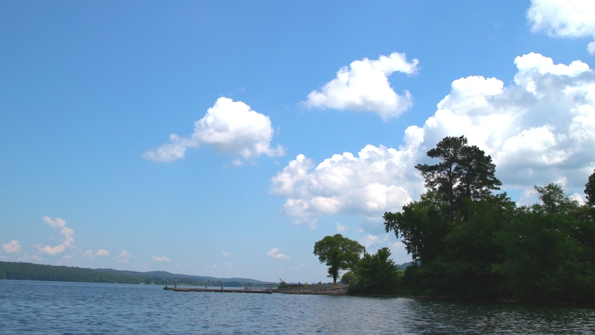 A beautiful clip of a blue sky with big white puffy clouds set over a serene lake with fishing dock jutting offshore.  Clip contains trees on the right foreground and rolling hills in background.