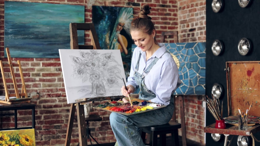 Portrait of female artist mixing color paints with art brush on the palette. Young beautiful painter at the studio preparing for painting a still life picture with sunflowers. Royalty-Free Stock Footage #1092066491